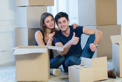 A young couple with light skin and dark hair, sitting on the floor amidst an array of opened cardboard moving boxes of various sizes in a well-lit, modern room with a large window and white curtains. The woman has straight hair, wearing a sleeveless navy dress, and is smiling while holding a small object. The man, with short hair, dressed in a dark blue T-shirt and jeans, is giving a thumbs-up gesture and has a cheerful expression. Behind them, the boxes are stacked and some are partially collapsed, indicating an ongoing packing or unpacking process typical of house removals in Balham or nearby areas within postcode SW12. The room features a light patterned carpet, with no furniture visible, emphasizing the focus on packing materials and the moving activity. The overall scene conveys a cheerful, organized approach to moving house, consistent with professional removals services offered by companies like Balham Man with Van.
