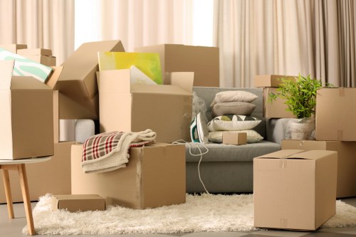 The image displays a cluttered living room scene during a house move, with numerous cardboard boxes of various sizes, some sealed and others open, containing household items. In the center, there is a light grey fabric sofa partially obscured by boxes, with folded blankets and a pile of cushions on top. To the left, a small wooden table supports a stack of flattened boxes. Several boxed belongings, including items wrapped in plastic and some with visible colorful papers or packaging, are scattered across a plush, cream-colored rug that covers the floor. Behind the sofa, beige curtains hang from a window, softly illuminated by natural daylight. To the right, more closed cardboard boxes are stacked, with a small potted green plant adding a touch of foliage. The scene exudes an active moving process within a residential interior, typical of Balham or South London homes, prepared for transition. The variety of packed items and open boxes highlight the packing stage prior to professional removals services from Balham Man with Van, reflecting a typical house relocation environment.
