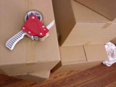 The image shows a close-up view of several large cardboard moving boxes stacked inside a residential space, likely in a hallway or room preparing for a house removal. The boxes are made of brown corrugated cardboard with visible creases and packing tape on some edges. In the foreground, there is a green and white tape dispenser with a red plastic handle, resting on top of one of the boxes, used for sealing packages during packing. The wooden flooring beneath the boxes features a natural, polished finish with visible wood grain patterns, indicative of a typical UK home environment. The lighting appears natural or softly diffused, illuminating the scene clearly. The overall scene captures the organized stage of packing in a relocation process, relevant to house removals services offered by companies like Balham Man with Van, operating in the Balham area or nearby postcode districts, reflecting the professional packing stage before transportation.