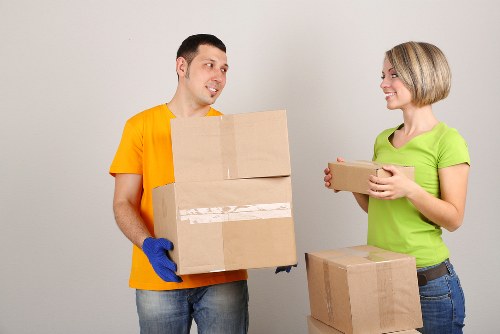 In an indoor residential environment, a male mover wearing a bright orange shirt, blue gloves, and jeans is holding a medium-sized cardboard box, facing a female colleague. The female, dressed in a light green T-shirt and jeans, is smiling and holding a smaller cardboard box, with another box placed on the floor next to her. The boxes are standard brown cardboard with visible tape securing their lids. The background features a plain, light grey wall with no additional furniture or items visible. The scene captures the process of packing or unpacking during a house removal, typical of professional removals services in the Balham area or nearby London locations. The lighting is neutral and evenly distributed, providing clear visibility of the items and the individuals involved, with no clutter or extraneous objects. The focus is on the interaction between the movers and their handling of the household belongings as part of a professional house removal operation carried out by Balham Man with Van, reflecting the clean and organized environment typical of residential moves in London.