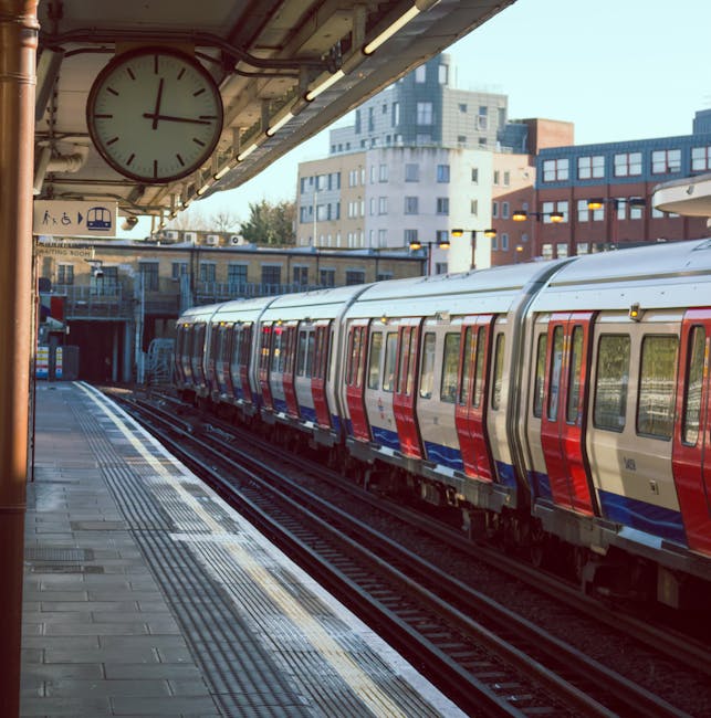 A woman with long hair, wearing a dark puffer jacket, stands on a train station platform, facing a train with sliding doors, with her hands in her pockets. In the background, other passengers are visi