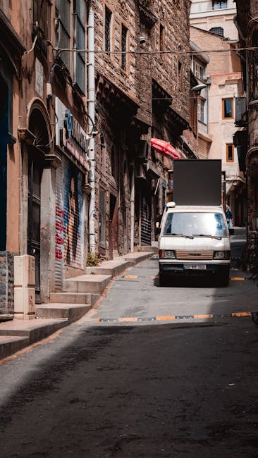A narrow urban street scene showing a small open area between tall buildings with dark facades on the left and lighter-colored residential or commercial structures on the right. The ground is paved wi